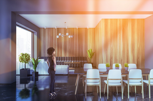 Young African American woman standing in modern dining room with gray and wooden walls, black marble floor and living room in background. Toned image