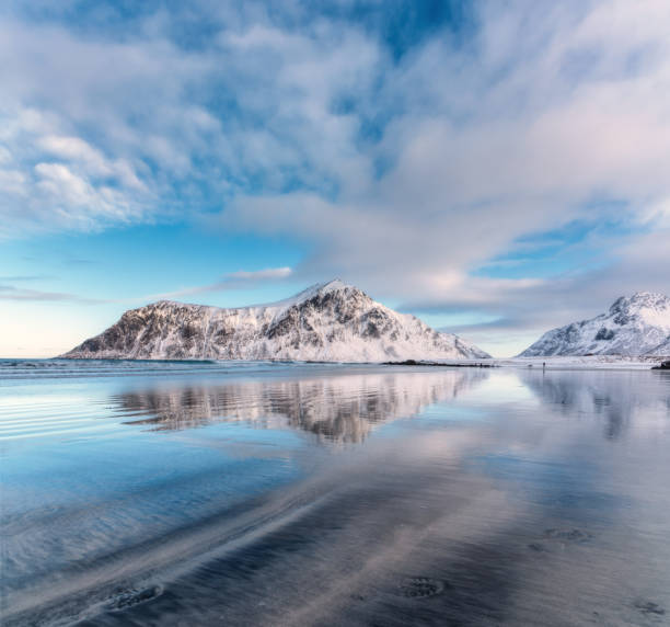 istock Snowy mountains and blue sky with clouds reflected in water in winter. Arctic sandy beach in Lofoten islands, Norway. Landscape with snow covered rocks, nordic sea coast, sand. Nature background 1288483742