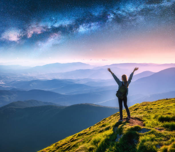 happy young woman on the mountain peak and arched milky way over mountains at night. landscape with girl, blue starry sky, milky way arch, green grass, hills in fog. space and galaxy. sky with stars - milky way galaxy space star imagens e fotografias de stock