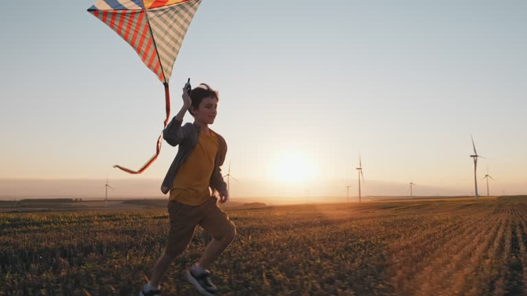 Happy boy runs launches bright kite into sky mown wheat field, playing with wind in field of an orange sunset on day lens flares wind turbines in summer slow motion. School break. Lifestyle. Childhood