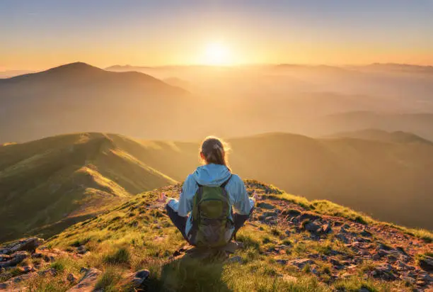 Photo of Young woman with backpack sitting on the mountain peak and beautiful mountains in fog at sunset in summer. Landscape with sporty girl, green forest, hills , sky, sunbeams. Travel and tourism. Yoga