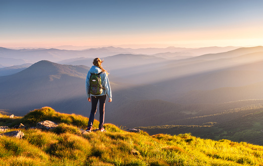 Beautiful mountains in fog and standing young woman with backpack on the peak at sunset in summer. Landscape with sporty girl, green grass, forest, hills , blue sky with sunbeams. Travel and tourism