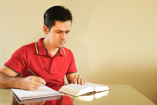 Adult man sitting at the table in his living room, with yellow background wall, reading the bible and writing notes on a paper