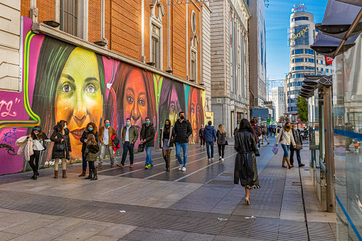 November 2020. Gran Via, Madrid, Spain. Street photography of masked people walking in the front of colorful painting of women's faces.