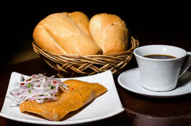 Photo of Peruvian breakfast, popular Peruvian tamale with onion sauce, breads and a cup of coffee on a table.