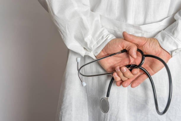 istock Doctor in white coat holds hands behind his back with stethoscope. Close up. 1288565415