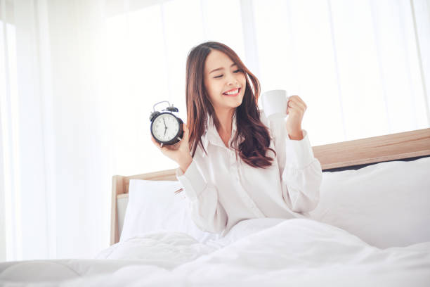 donna che tiene la sveglia e una tazza di caffè sul letto al mattino. - yawning women drink coffee cup foto e immagini stock