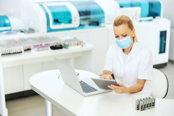 istock Attractive blond lab assistant in coat with sterile surgical mask on sitting in laboratory, entering test results on tablet. Research for cure for covid 19 concept. 1288571845