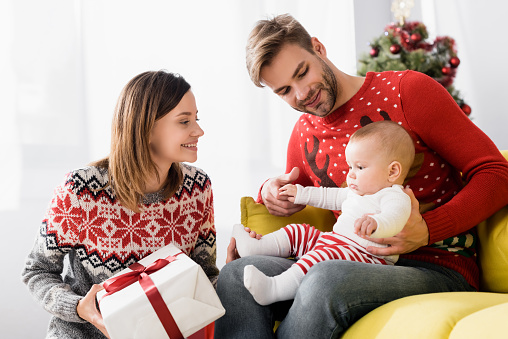 happy mother holding wrapped christmas present near husband and baby son