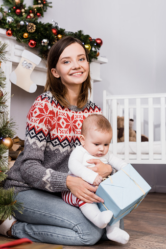 cheerful woman holding present near baby boy in decorated apartment