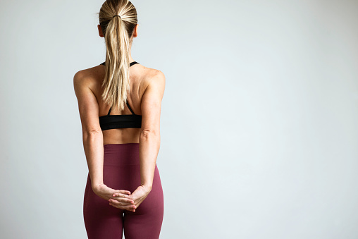 Back view of caucasian woman wearing sportswear is standing with her arms behind  in front of white wall .Note : Image is not body shape retouched.