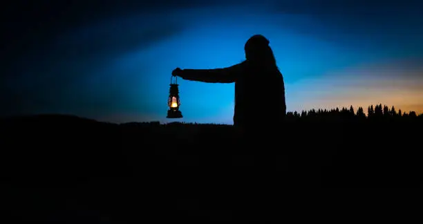 Photo of Silhouette of a woman looking into the last light of the day by a lake in the wild, holding a lit vintage kerosene lamp.