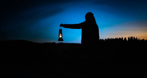 silhouette of a woman looking into the last light of the day by a lake in the wild, holding a lit vintage kerosene lamp. - kerosene imagens e fotografias de stock