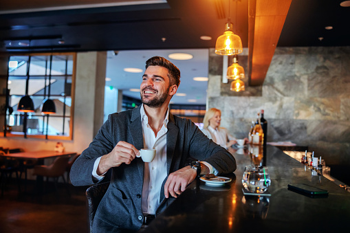 Smiling middle-aged man in formal wear sitting in the bar and drinking his morning coffee.