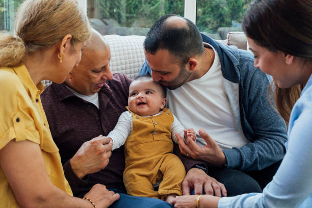 Multi Gen Family Meeting Baby Three generation family sitting together in their home talking and laughing together. There is a baby boy sitting in the middle of them and the father is kissing his head. indian couple kiss stock pictures, royalty-free photos & images
