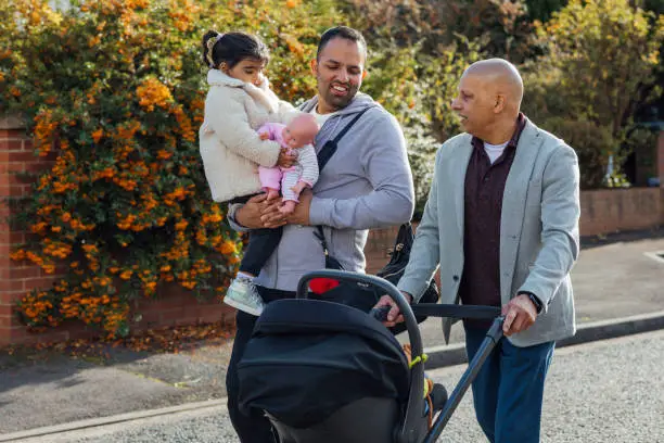 Multi generation family on a walk together.  There is a father, son and his sons children. The grandfather is pushing a pushchair while his son is holding his daughter who is carrying a toy doll.