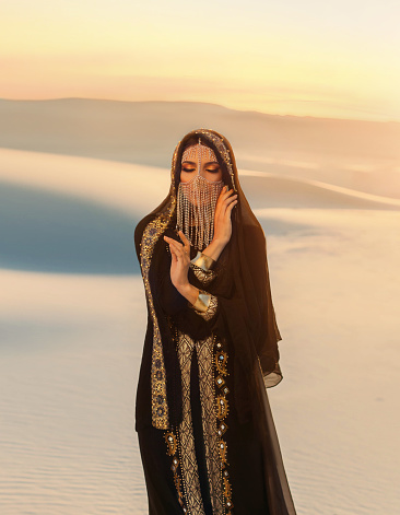 Beautiful mysterious arab woman Queen in black muslim dress. head with silk handkerchief, golden chains hides face. A Fantasy Girl oriental princess walks in desert. art background yellow sand sunset