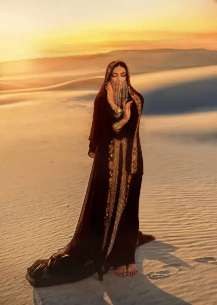 Photo of A mysterious woman in a black long dress stands in the desert. Luxurious clothes, gold accessories hide the face. Asian beauty fashion model. Creative makeup. Sand dunes background, orange sunset