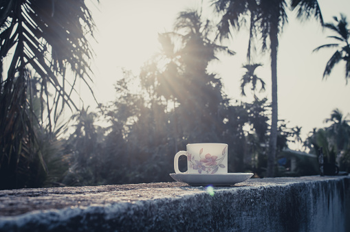 Coffee cup in sunset sunlight. Summer fresh cool look. White coffee cup on saucer for hot drink on roof beam of a residential building with bokeh city in the background.