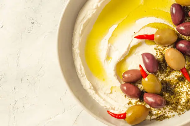 Photo of Popular middle eastern appetizer labneh or labaneh, soft white goat milk cheese with olive oil, hyssop or zaatar, olives on grey table, top view ,flat lay. Close up image.