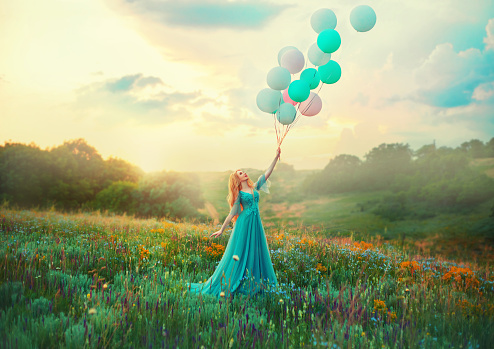 Happy woman walks in blooming valley. Fantasy girl princess holding in hand ball air balloon. long blue tulle fashion dress. Sunset sky, magic haze fog, flowers green meadow. blonde hair. Smiling face