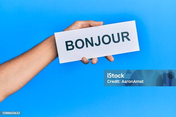 Hand Of Hispanic Man Holding French Greeting Bonjour Word Paper Over Isolated Blue Background Stock Photo - Download Image Now