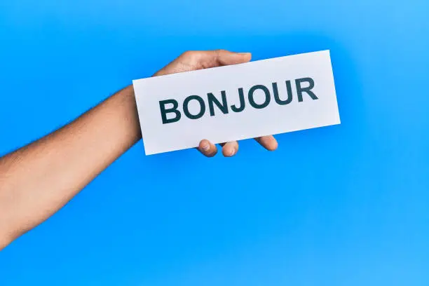 Hand of hispanic man holding french greeting bonjour word paper over isolated blue background.