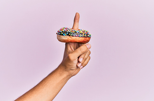 Finger of hispanic man holding chocolate donut over isolated pink background.