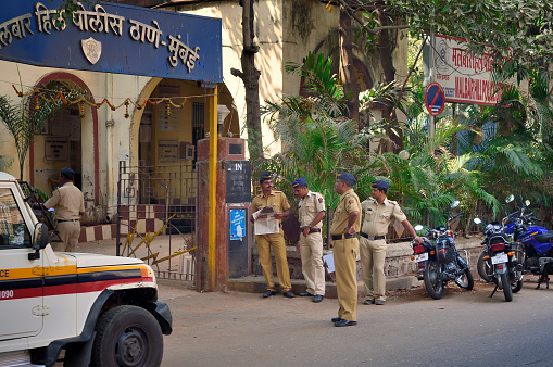 Four policemen wearing military style overseas caps  stand outside the Malbar Hill police station in Mumbai, India.  Two are reading a newspaper, one leans on motorcycle while the fourth appears to be a senior officer standing with a clipboard in his hand in April, 2012,