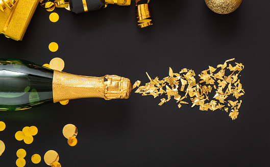Golden bottle of champagne spills out gold sparkles in frame of gold festive Christmas decor confetti balls gifts on black background. Flat lay New Year eve celebration dark backdrop. Long web banner.