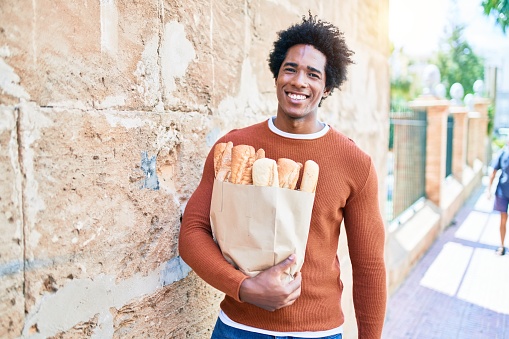 Young handsome african american man smiling happy. Standing with smile on face holding paper bag with bread walking at town street.