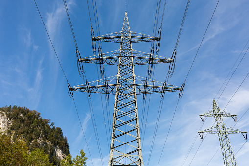 Kochelsee, Germany - Oct 1, 2020: Overhead power line with blue sky and an upper bavarian mountain. Providing electricity from a hydroelectric power plant.