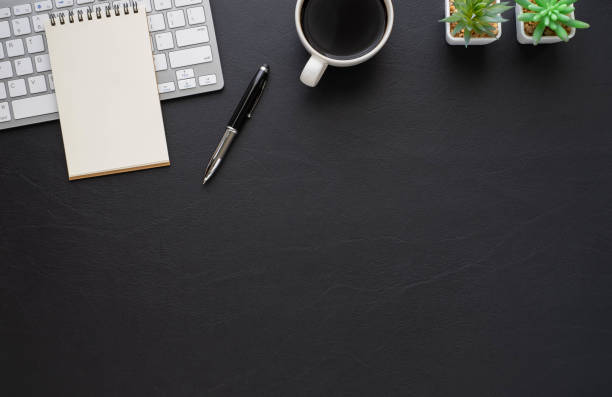 istock Top view above of Black leather office desk table with keyboard, notebook and coffee cup with equipment other office supplies. Business and finance concept. Workplace, Flat lay with blank copy space. 1288613367