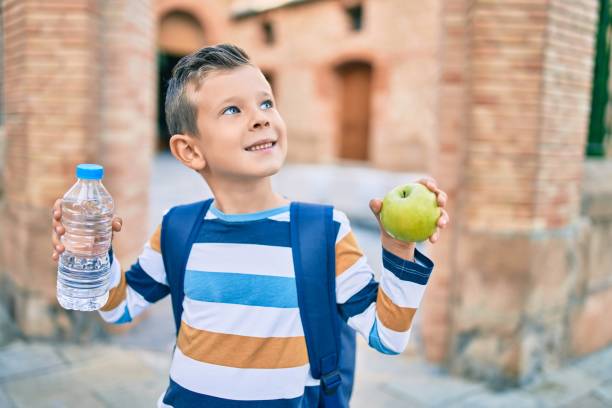 garçon caucasien adorable d’étudiant souriant heureux retenant la pomme verte et la bouteille d’eau à la ville. - people eating walking fun photos et images de collection