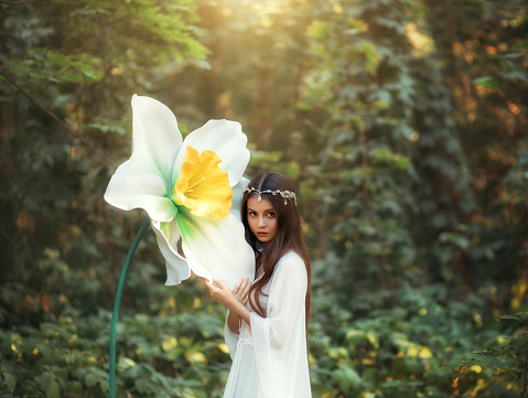 Portrait of a beautiful girl elf with long hair stands in a fantasy forest and hugs a huge daffodil flower, green trees. A woman in a white vintage dress and a silver diadem. Innocent attractive face.