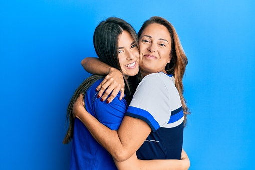 Beautiful hispanic mother and daughter smiling happy hugging over isolated blue background.
