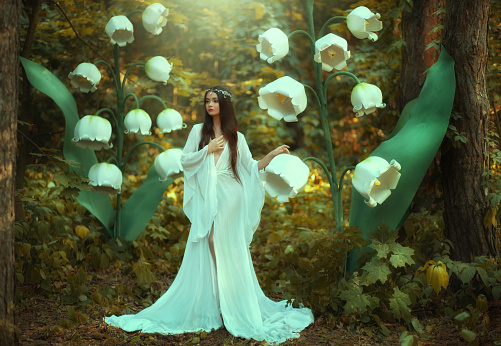 Beautiful fantasy woman princess elf in long white dress walks in fairy forest with large flowers lilies of valley. Queen girl in silver diadem. Silk vintage outfit with wide sleeves. Green trees.