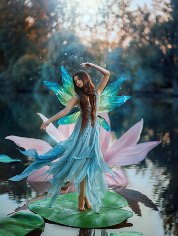 Beautiful young fantasy woman in the image of a river fairy dances on a water lily flower. A long silk dress flies in the wind, butterfly wings glisten. Background evening dark nature, blue lake
