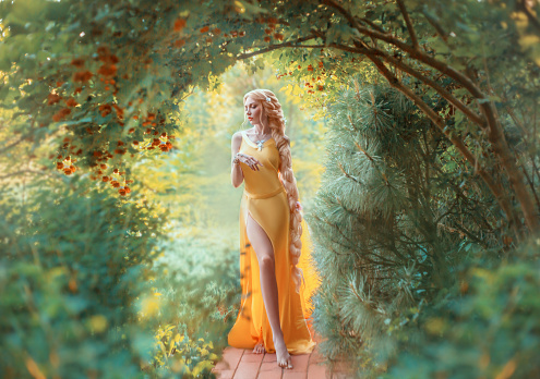 Beautiful young blond woman with very long hair that is braided. The girl is dressed in a seductive yellow dress with a slit on the leg. Fashion model posing against the backdrop of an autumn garden