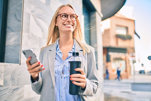 Young blonde businesswoman using smartphone and holding bottle of water at the city.
