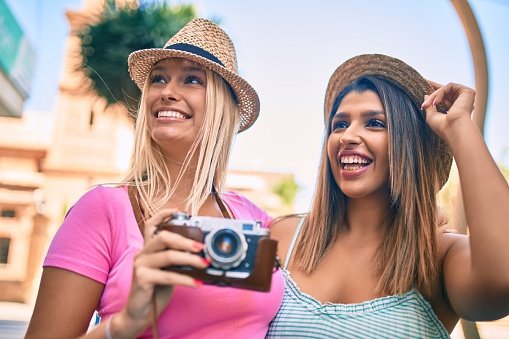 Two beautiful and young girl friends together having fun at the city standing with vintage camera