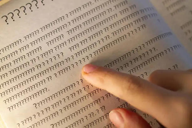 Photo of A person leads a finger on the lines in the book, but instead of letters only question marks on the page in the textbook