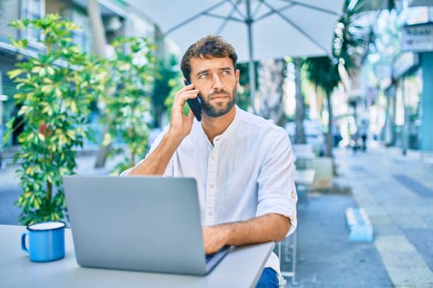 istock Handsome man with beard wearing casual white shirt on a sunny day working using laptop and speaking on the phone at cafeteria 1288622987