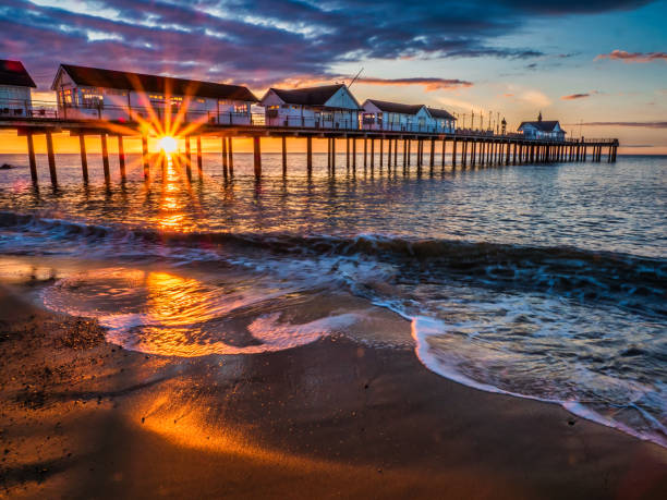Southwold pier at sunrise with waves lapping up sandy beach and sun shining through structure, Southwold, Suffolk, England, Britain