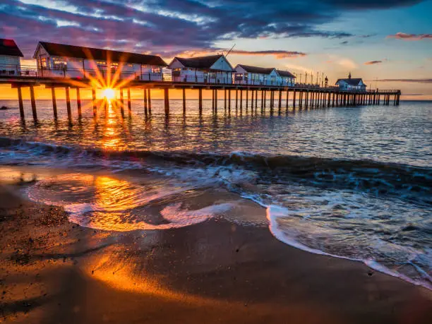 Photo of Southwold pier at sunrise with waves lapping up sandy beach and sun shining through structure, Southwold, Suffolk, England, Britain