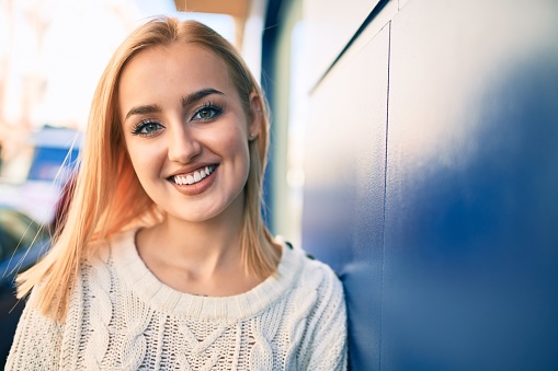 Young blonde girl smiling happy leaning on the wall at the city.