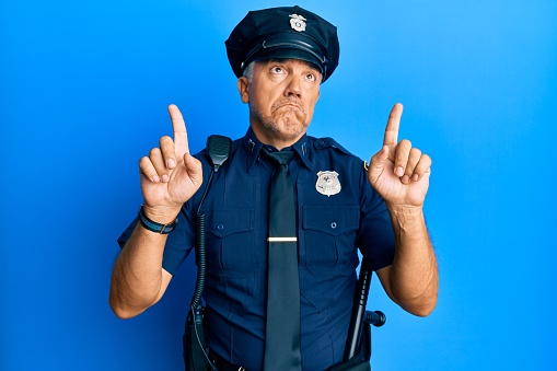 Handsome middle age mature man wearing police uniform pointing up looking sad and upset, indicating direction with fingers, unhappy and depressed.