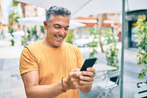 hombre de pelo gris de la edad media sonriendo feliz usando el teléfono inteligente caminando en la calle de la ciudad. - person on phone fotografías e imágenes de stock