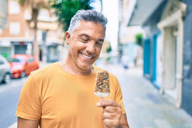 mittleres alter grauhaariger mann lächelnd glücklich essen eis auf der straße der stadt. - people eating walking fun stock-fotos und bilder