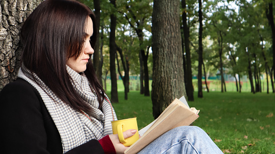 A woman sits under a tree and reads her favorite book while drinking coffee or tea from a yellow cup in a city park on green grass on a pleasant sunny day. Vacation, education and study concept